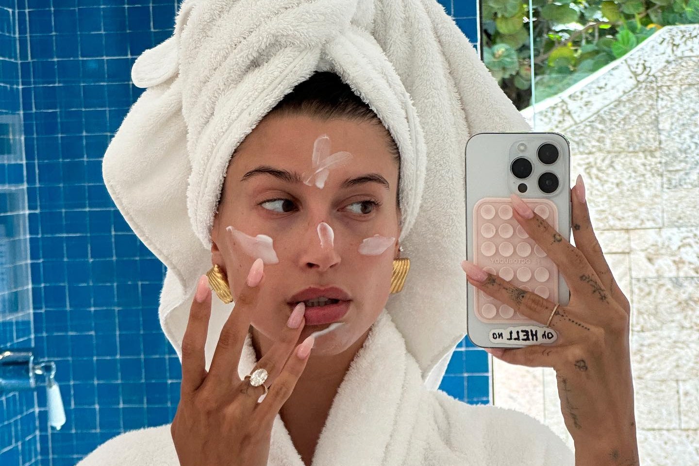 Hailey Bieber takes a mirror selfie in a bathroom with moisturizer applied to her face while wearing a towel wrapped...