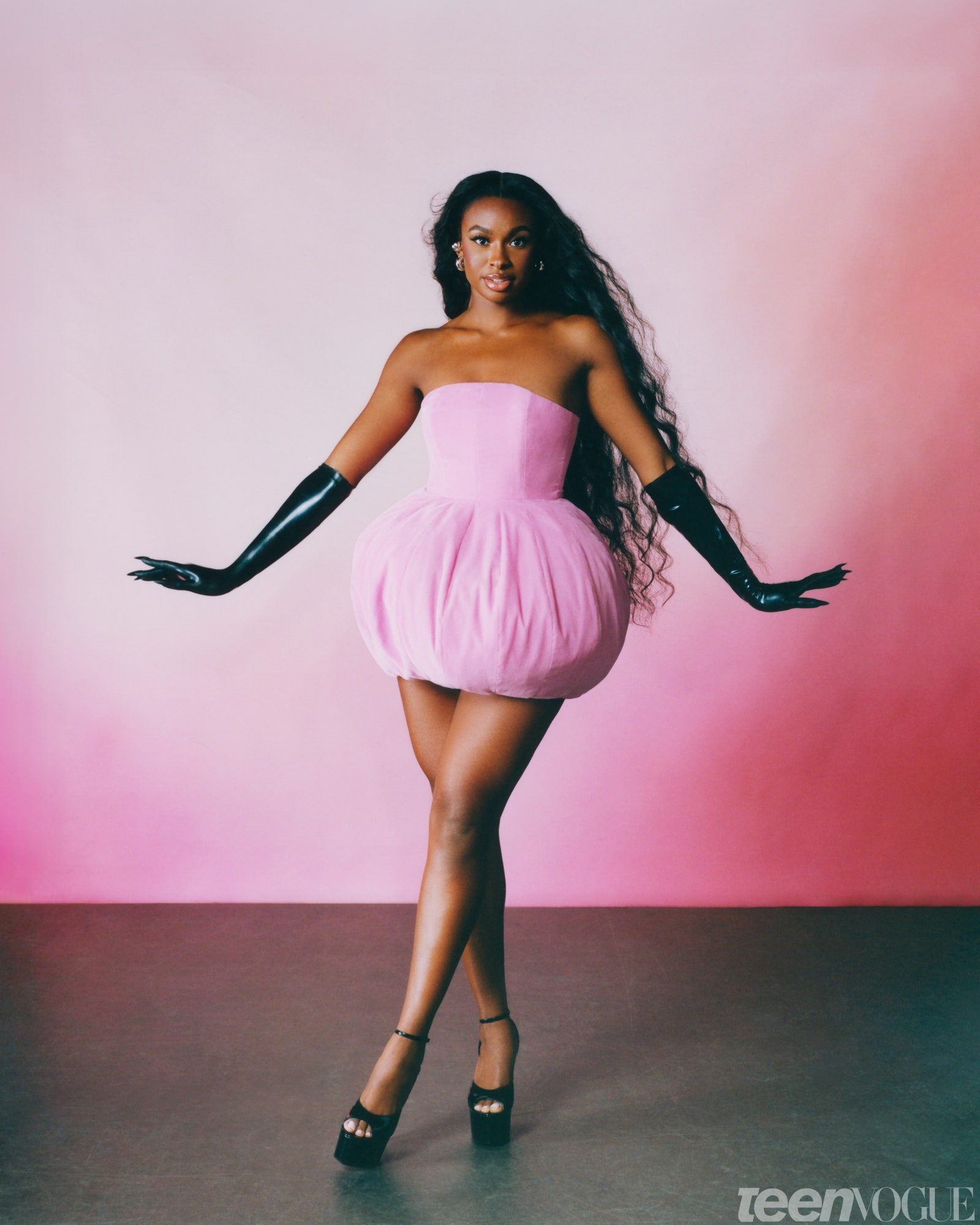 Coco Jones standing against a pink background wearing a pink dress and black latex gloves