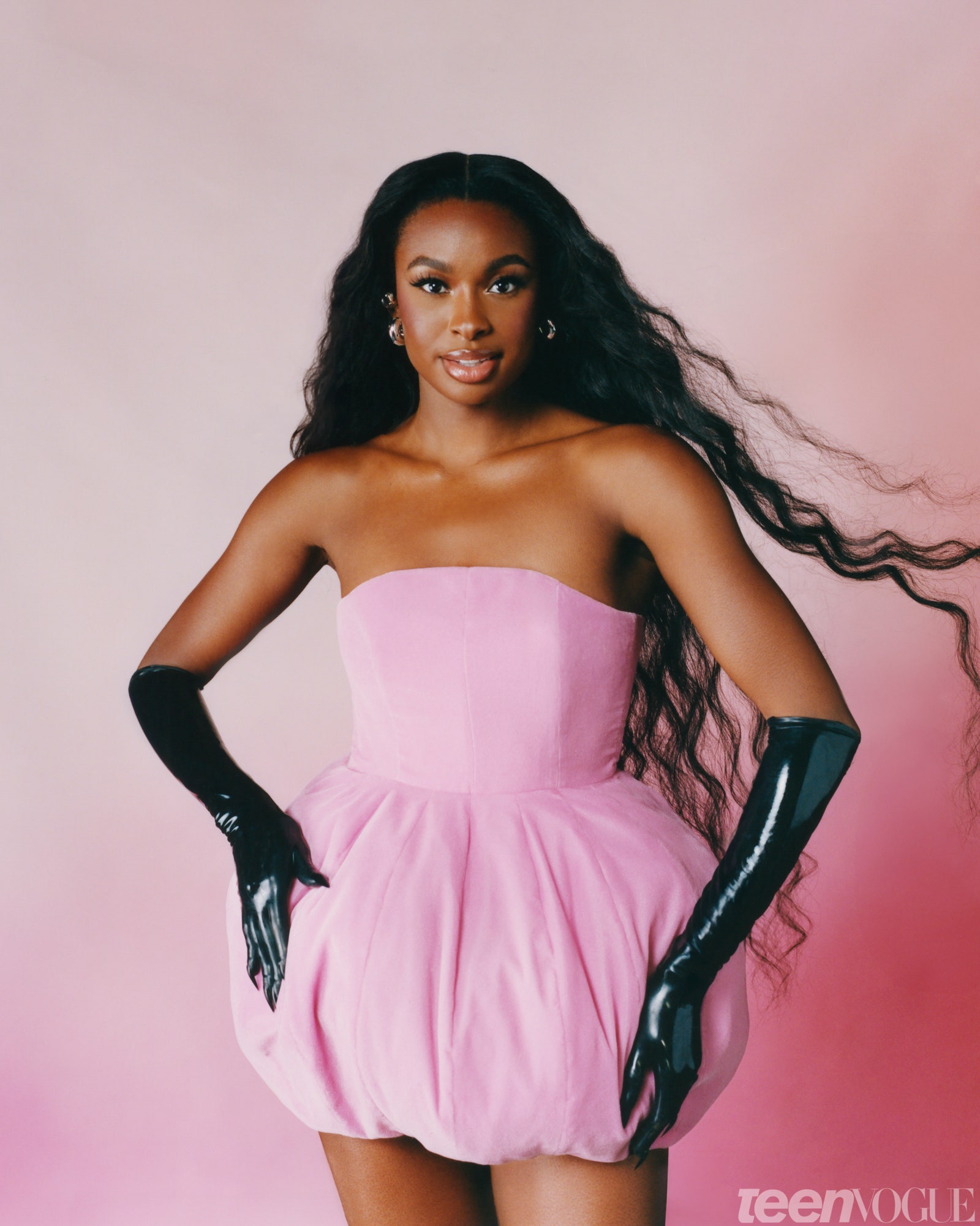 Coco Jones standing against a pink background wearing a pink dress and black latex gloves