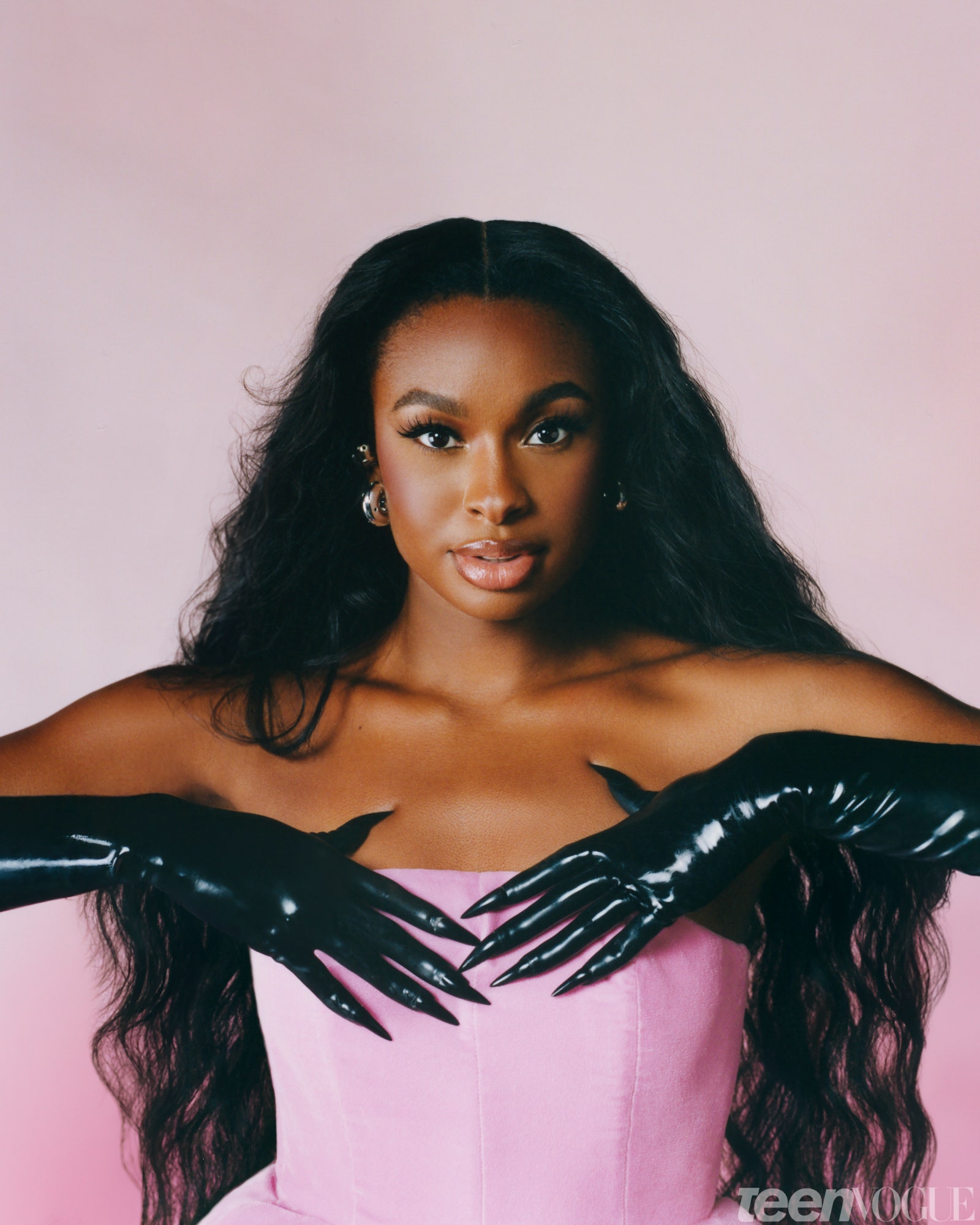 Coco Jones against a pink background wearing a pink dress and black latex gloves